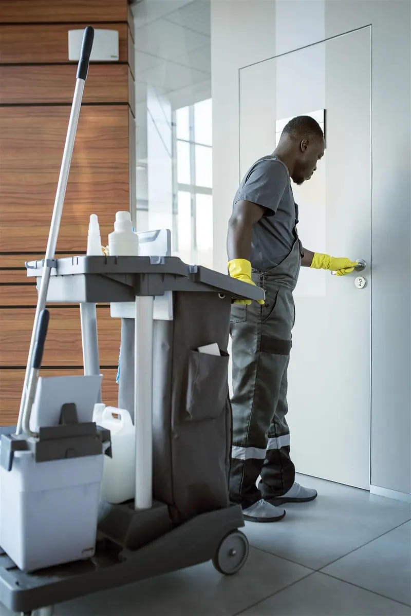 Cleaner in yellow gloves beside a supplies trolley, opening an office door